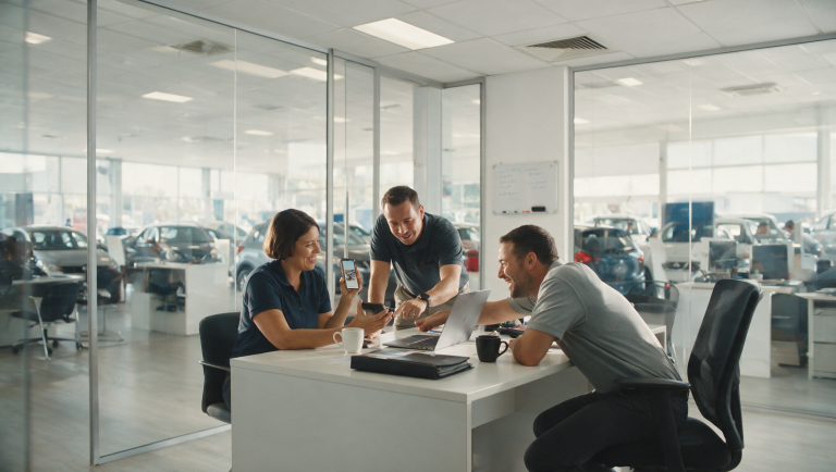 Three salespeople in a South African dealership sales office looking at a social media post on a phone together, showroom visible behind them